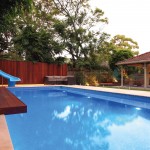 A modern blue pool next to a house with outdoor furniture and a water slide